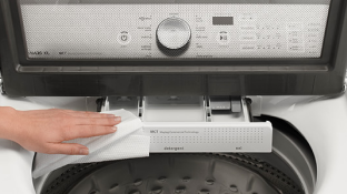 Une laveuse avec la porte ouverte. Une main nettoie l'ouverture de la laveuse avec une lingette blanche. Par le hublot de la porte, on peut voir le panneau de commande