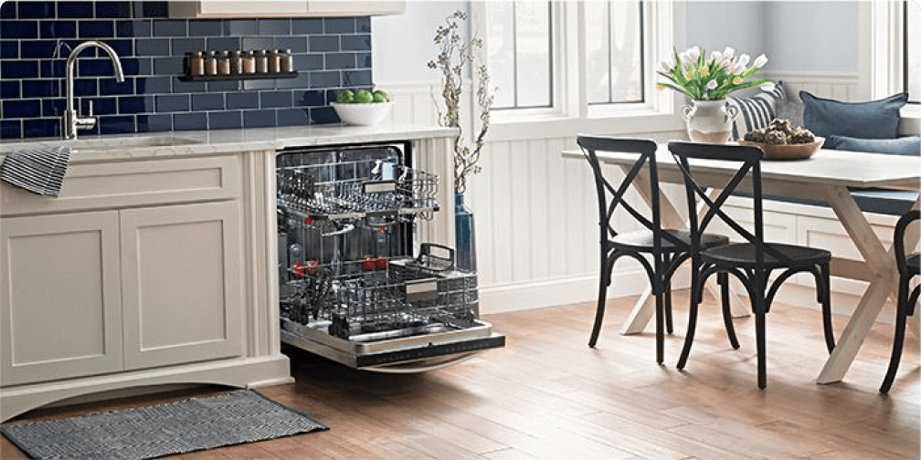 In a kitchen with wood floors, white cabinetry and a dark blue backsplash is a table with three chairs and a opened dishwasher with the racks pulled out. On the marble countertops is a bowl of green apples and on the wall is a shelf lined with spices.