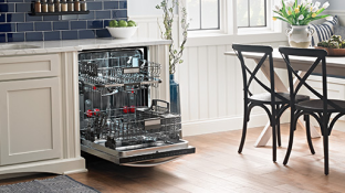 Une cuisine avec un lave-vaisselle ouvert avec les paniers vides sortis. Le lave-vaisselle est entouré d'armoires blanches, d'une table avec deux chaises et d'un plancher en bois. Sur le comptoir au-dessus du lave-vaisselle se trouve un bol de pommes vertes et une étagère à épices est accrochée au mur