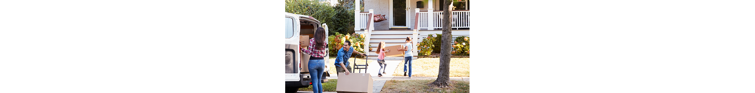 A family moving into a house. Two kids carry a box toward the house, a woman is about to remove a box from the back of a van filled with boxes and a man tilts a dolly with a box on it.