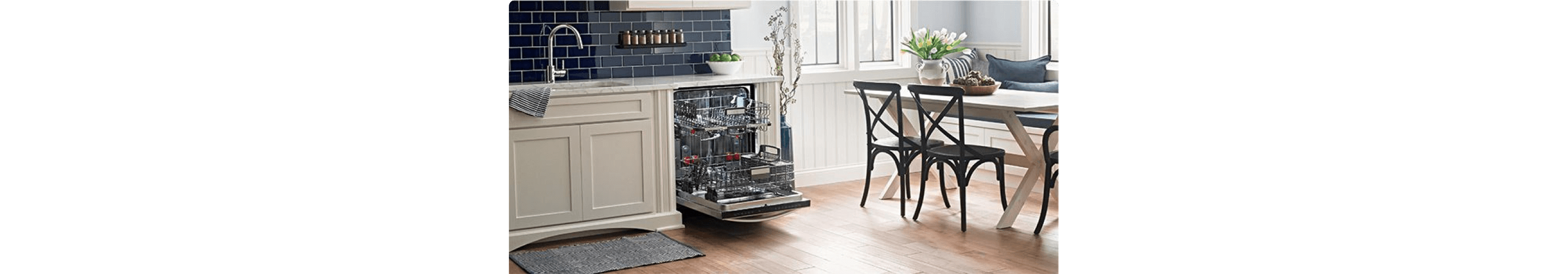 In a kitchen with wood floors, white cabinetry and a dark blue backsplash is a table with three chairs and a opened dishwasher with the racks pulled out. On the marble countertops is a bowl of green apples and on the wall is a shelf lined with spices.