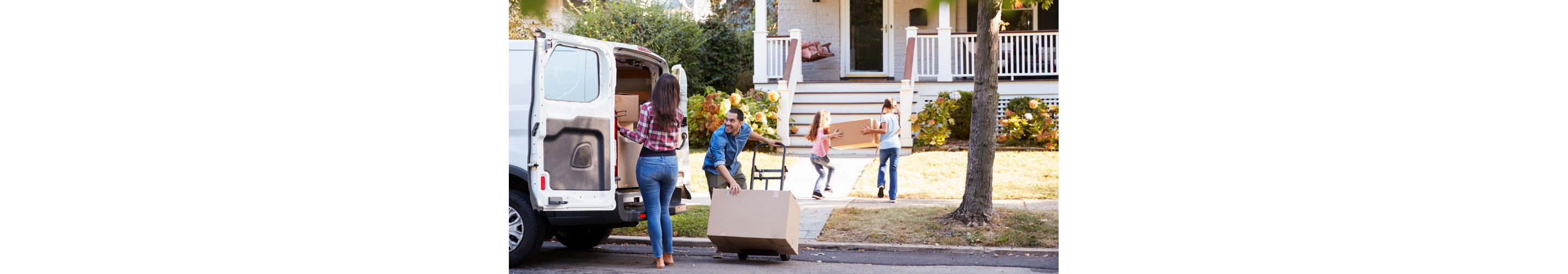 A family moving into a house. Two kids carry a box toward the house, a woman is about to remove a box from the back of a van filled with boxes and a man tilts a dolly with a box on it